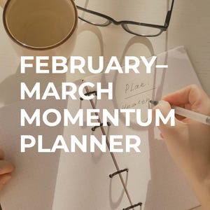 May include: A light-colored ceramic mug, eyeglasses, and a planner are on a white surface. The planner is open, with the text "FEBRUARY-MARCH MOMENTUM PLANNER" and a hand writing on the page. The planner is bound with black rings and pink string.