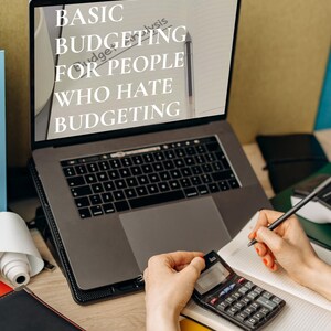May include: A laptop displays the text "BASIC BUDGETING FOR PEOPLE WHO HATE BUDGETING." A person uses a calculator and writes in a notebook. Other items include a pen, a roll of paper, and a black notebook.