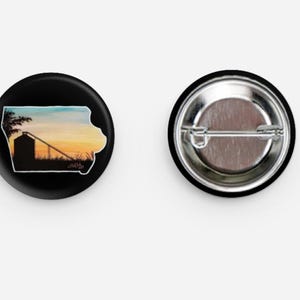 May include: Two round black buttons. One button features a silhouette of Iowa's outline with a sunset scene inside. The other button shows the pin back. The Iowa button has a white border and a sunset with a silo.
