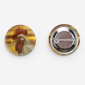 May include: Two round buttons. One button features a detailed illustration of a rooster's head with red comb and wattle, gray feathers, and a yellow beak, set against a golden background. The other button shows the metal back.