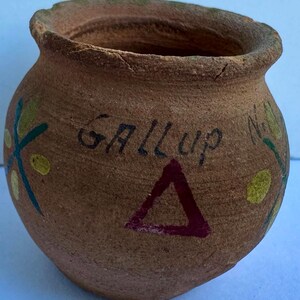 May include: A small, handmade terracotta pot with a slightly flared rim. The pot features the word "GALLUP" in black, a red triangle, and painted floral designs in yellow, green, and blue. The pot has a rustic, aged appearance.