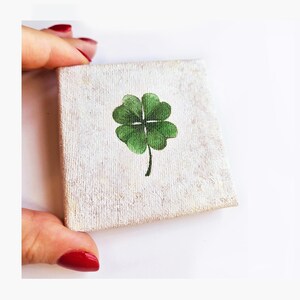 May include: A small, square canvas with a hand-painted green four-leaf clover. The canvas has a textured, off-white background. The clover is centered and has detailed green leaves and a black stem.