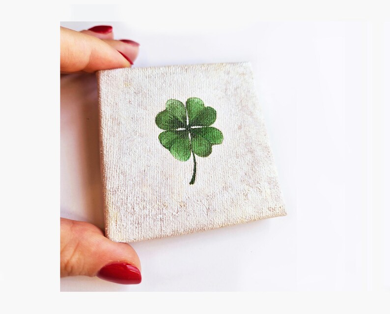 May include: A small, square canvas with a hand-painted green four-leaf clover. The canvas has a textured, off-white background. The clover is centered and has detailed green leaves and a black stem.