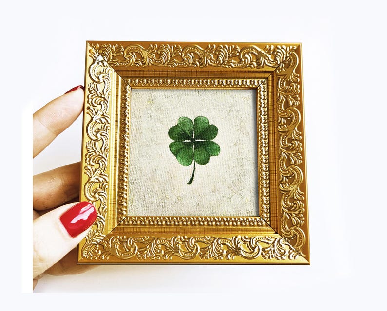 May include: A small, square, gold-colored picture frame with ornate detailing. Inside the frame is a canvas with a green four-leaf clover. The frame is held by a hand with red nail polish.