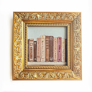 Puede incluir: Una obra de arte enmarcada que representa una fila de libros en tonos marrones, rosas y beige. El marco es dorado con un diseño elaborado y ondulado. El fondo es azul claro. La obra es de forma cuadrada.