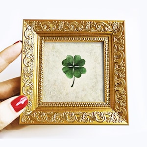 May include: A small, square, gold-coloured picture frame with ornate detailing. The frame holds a small artwork featuring a green four-leaf clover on a textured, off-white background. The frame is held by a hand with red nail polish.