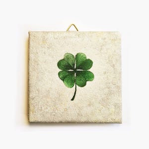 May include: Square canvas art featuring a green four-leaf clover on a textured, off-white background. A small gold-colored hook is attached at the top for hanging. The artwork has a rustic, vintage aesthetic.