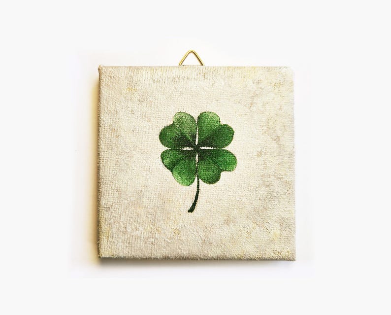 May include: Square canvas art featuring a green four-leaf clover on a textured, off-white background. A small gold-colored hook is attached at the top for hanging. The artwork has a rustic, vintage aesthetic.