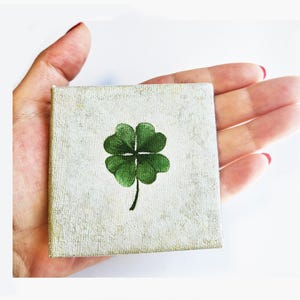 May include: A small square canvas with a hand-painted green four-leaf clover on a textured, off-white background. The artwork is held in a hand, showcasing the miniature size of the lucky charm.