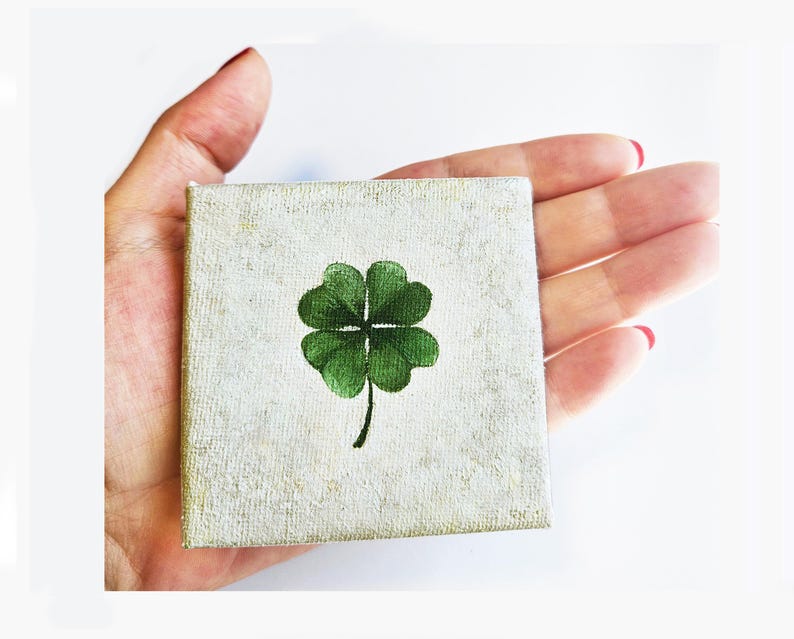 May include: A small square canvas with a hand-painted green four-leaf clover on a textured, off-white background. The artwork is held in a hand, showcasing the miniature size of the lucky charm.