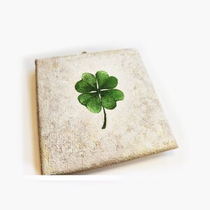 May include: A square canvas with a textured, off-white background features a hand-painted green four-leaf clover. The clover is centered on the canvas, with a dark green stem extending downwards. The artwork is a symbol of good luck.