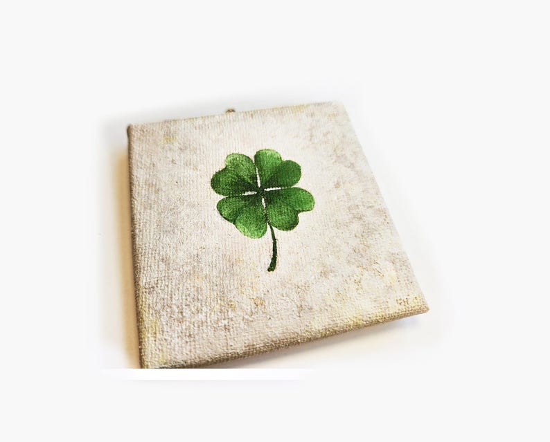 May include: A square canvas with a textured, off-white background features a hand-painted green four-leaf clover. The clover is centered on the canvas, with a dark green stem extending downwards. The artwork is a symbol of good luck.