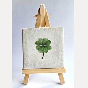 May include: A small, square canvas painting of a green four-leaf clover on a miniature wooden easel. The clover is painted with detailed shading, and the canvas has a textured, off-white surface. The easel is made of light-colored wood.