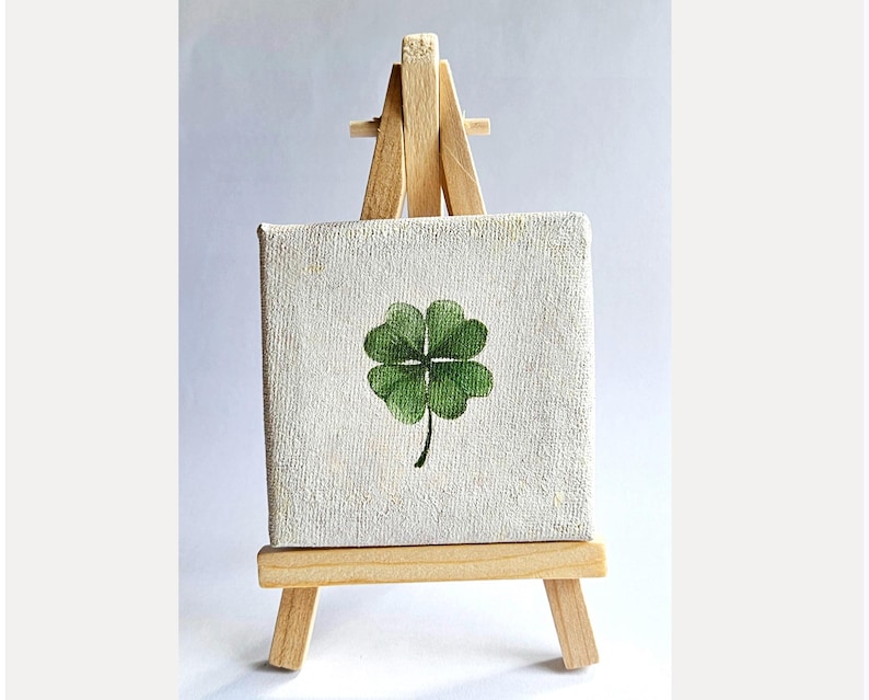 May include: A small, square canvas painting of a green four-leaf clover on a miniature wooden easel. The clover is painted with detailed shading, and the canvas has a textured, off-white surface. The easel is made of light-colored wood.