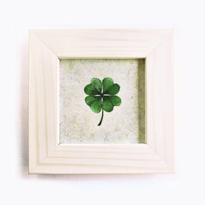 May include: A framed artwork featuring a green four-leaf clover on a textured background. The clover is centered within a square, light-colored wooden frame. The artwork is a symbol of good luck and is displayed against a white backdrop.