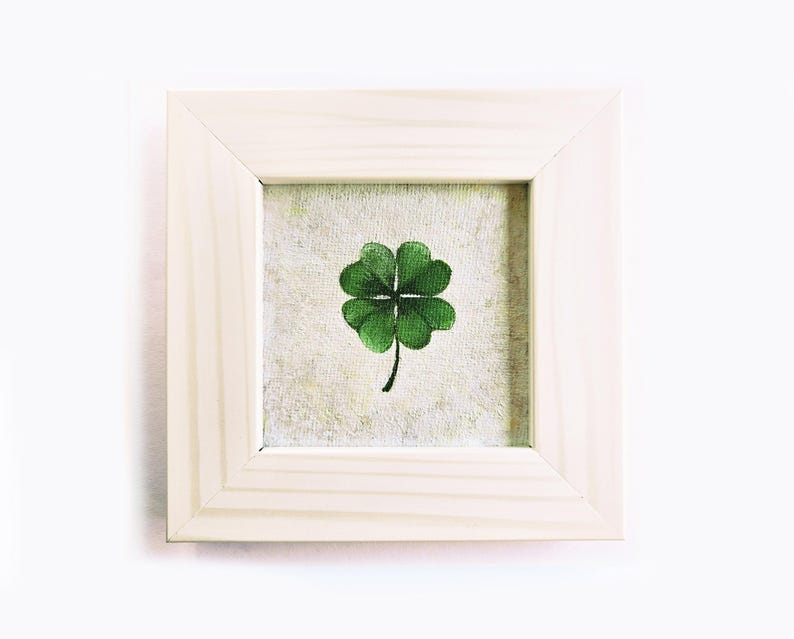 May include: A framed artwork featuring a green four-leaf clover on a textured background. The clover is centered within a square, light-colored wooden frame. The artwork is a symbol of good luck and is displayed against a white backdrop.