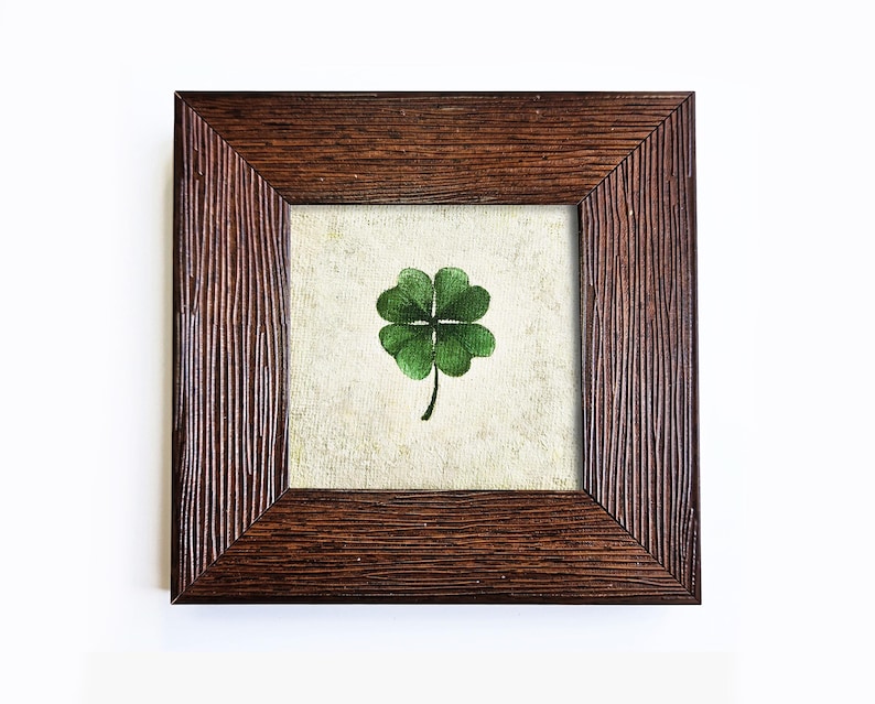 May include: A framed artwork featuring a green four-leaf clover on a cream-colored background. The artwork is encased in a square, dark brown wooden frame with a textured, vertical grain pattern. The image is set against a white backdrop.
