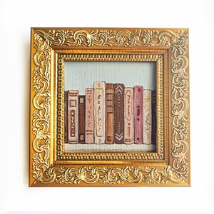 May include: A square embroidery piece featuring a row of books with spines in shades of brown, pink, and cream, set against a light blue background. The artwork is encased in a thick, ornate, metallic gold frame with intricate scrollwork patterns.