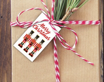 May include: A white gift tag with the text "Merry Christmas" in red cursive lettering. Three toy soldier illustrations are featured on the tag. The tag is tied with red and white twine to a brown gift box.