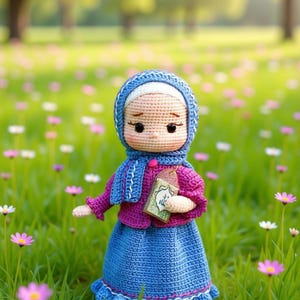 Puede incluir: Muñeca de ganchillo hecha a mano con hiyab, bufanda y falda azules, con un suéter rosa. La muñeca sostiene un pequeño libro con detalles verdes y dorados, de pie en un campo de hierba verde y flores rosas y blancas.