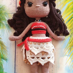 Może przedstawiać: Szydełkowa lalka o brązowej karnacji, ciemnobrązowych włosach i czerwono-białym stroju. Lalka ma kwiat we włosach i jest ustawiona na tropikalnym tle. Spódnica lalki jest biała z dekoracyjnym wzorem.