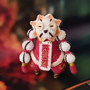 May include: A handmade crochet doll with a unique design. The doll features a white head with button eyes, a red and white outfit, and gold accents. The doll has a distinctive, whimsical appearance, perfect for collectors or as a decorative item.