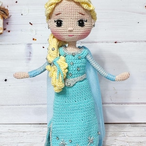 Puede incluir: Una muñeca de crochet con el pelo largo y rubio, vestida con un vestido azul claro con detalles plateados y una capa a juego. La muñeca tiene una expresión dulce y ojos grandes y oscuros.