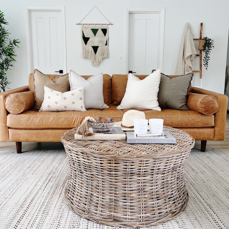 May include: A wicker coffee table with a woven design sits in front of a brown leather sofa with multiple throw pillows. The sofa is in a living room with white walls and a macrame wall hanging.