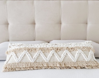 Puede incluir: Un cojín lumbar tejido blanco y beige con un borde de flecos. El cojín tiene un patrón de chevron y está sentado en una cama blanca.