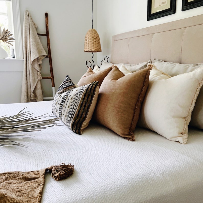 May include: A white bed with a white, brown, and black patterned throw pillow, a brown throw pillow, and a white throw pillow with fringe. The bed has a white duvet cover and a brown throw blanket at the foot of the bed.