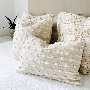May include: Three cream-coloured throw pillows with a textured woven design. The pillows have a raised, bumpy surface and are arranged on a white bed.