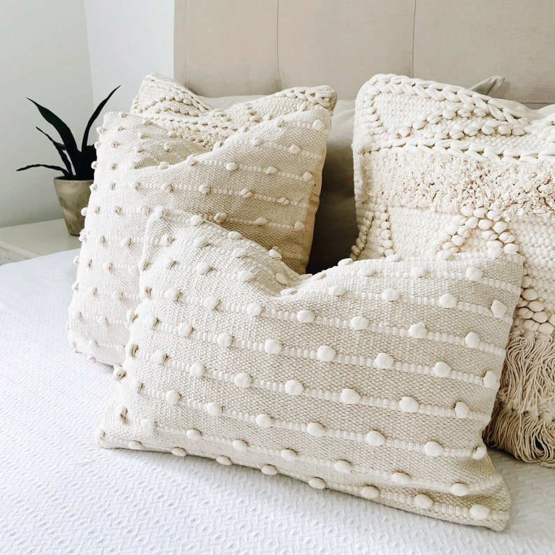 May include: Three cream-coloured throw pillows with a textured woven design. The pillows have a raised, bumpy surface and are arranged on a white bed.