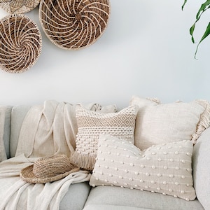 May include: A white couch with three pillows, two are white with a textured pattern and one is beige with a woven pattern. A straw hat is on the couch. Three woven baskets hang on the wall behind the couch.