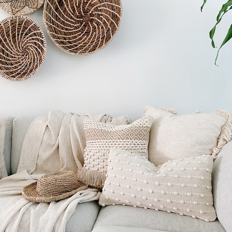 May include: A white couch with three pillows, two are white with a textured pattern and one is beige with a woven pattern. A straw hat is on the couch. Three woven baskets hang on the wall behind the couch.