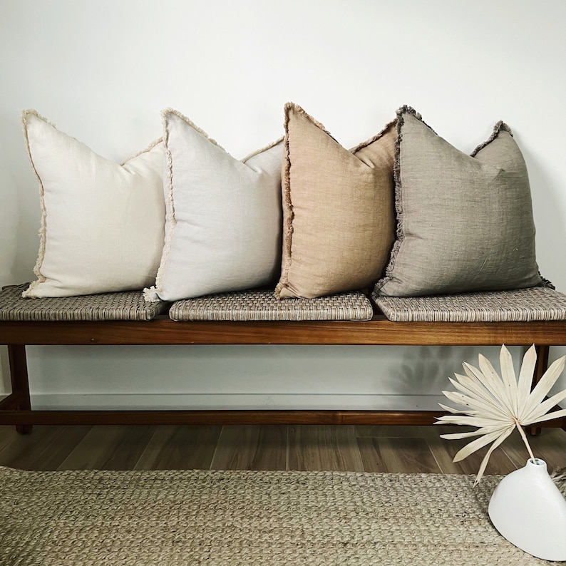 May include: Four decorative throw pillows with fringed edges, arranged on a wooden bench with a woven seat. The pillows are in shades of beige, cream, and light brown.