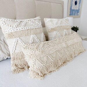 May include: Three cream-colored throw pillows with a woven texture and fringe trim. The pillows are arranged on a white bed with a cream-colored headboard.