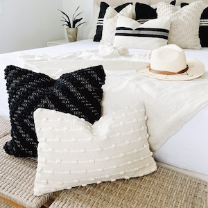 May include: A white bed with black and white pillows, a white throw blanket, and a straw hat. The bed is made with a white duvet cover and has a white headboard. The pillows are all different sizes and shapes, and some have a textured surface. The throw blanket is folded at the foot of the bed, and the straw hat is sitting on top of it.
