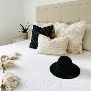 May include: A white bed with a black and white patterned throw blanket and three pillows. The pillows are white with black trim, white with a textured surface, and white with a fringe trim. A black hat is on the bed.