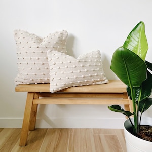 May include: Two cream-colored throw pillows with a textured, dotted design are placed on a light wood bench. A green plant with large leaves is in a white pot to the right of the bench.