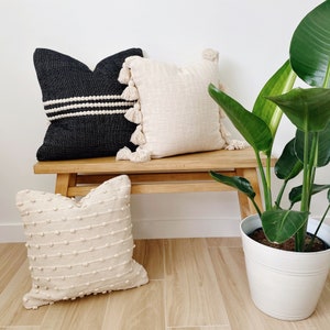 May include: Three decorative throw pillows on a wooden bench with a potted plant in the background. The pillows are made of different materials and have different textures. The bench is made of light wood and has a natural finish. The plant is a large green plant with large leaves.
