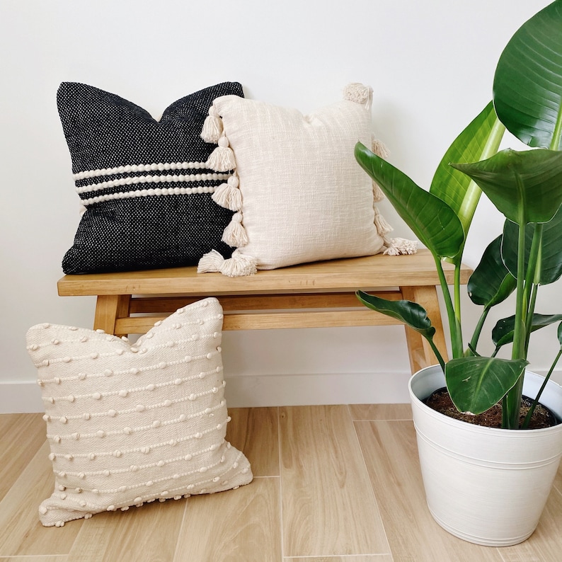 May include: Three decorative throw pillows on a wooden bench with a potted plant in the background. The pillows are made of different materials and have different textures. The bench is made of light wood and has a natural finish. The plant is a large green plant with large leaves.