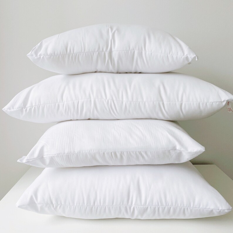May include: Four white pillows stacked on top of each other. The pillows are all made of a soft, white fabric and have a smooth, even surface.