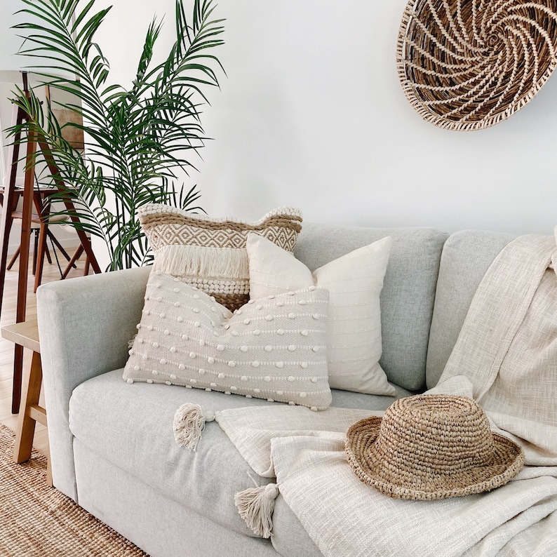 May include: A white couch with a variety of throw pillows, including a cream-colored pillow with a textured pattern, a tan pillow with a geometric pattern, and a white pillow with a fringe trim. A woven straw hat sits on the couch, and a woven basket hangs on the wall behind the couch.