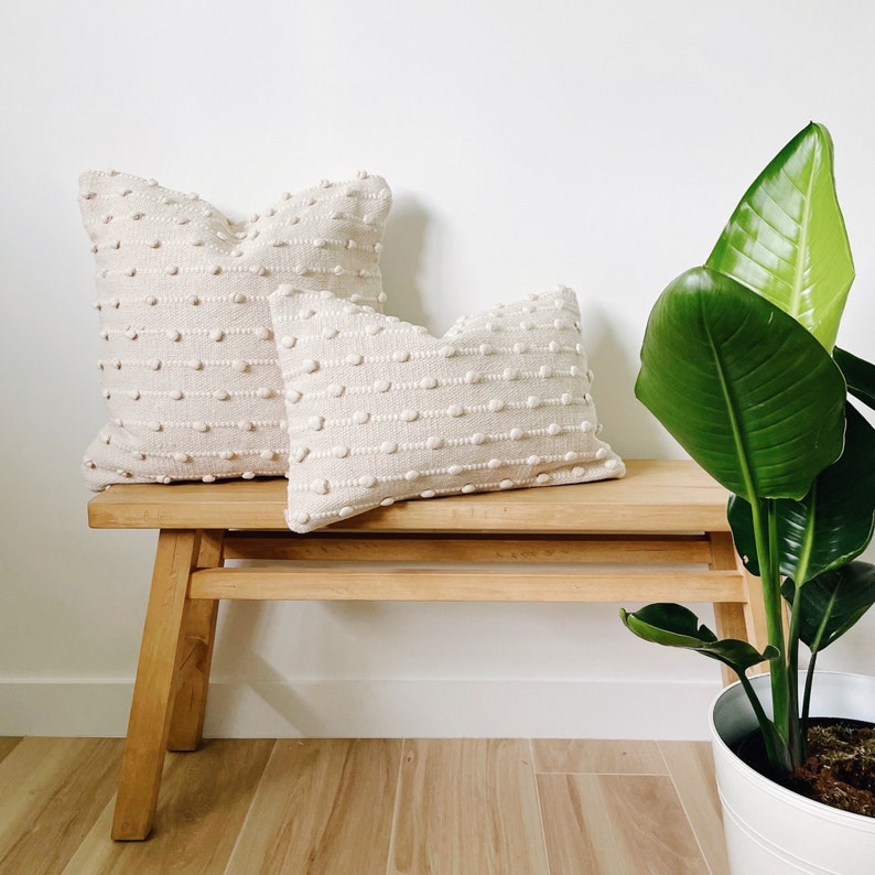 May include: Two off-white textured throw pillows with a subtle raised dot pattern are placed on a light wood bench. A large green leafy plant is in a white pot to the right of the bench.