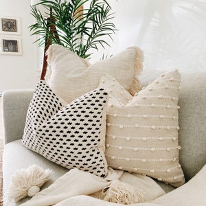 May include: A white sofa with a throw blanket and four cushions. Two of the cushions are white with a textured design, one is white with a fringe trim, and one is black and white with a geometric pattern.