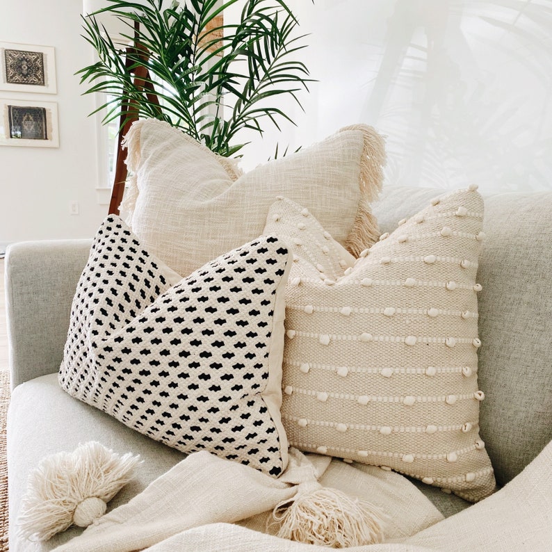 May include: A white sofa with a throw blanket and four cushions. Two of the cushions are white with a textured design, one is white with a fringe trim, and one is black and white with a geometric pattern.