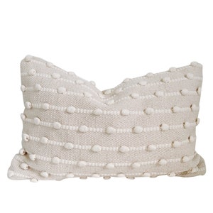 May include: A cream-coloured throw pillow with a textured woven design featuring small white pom-poms.