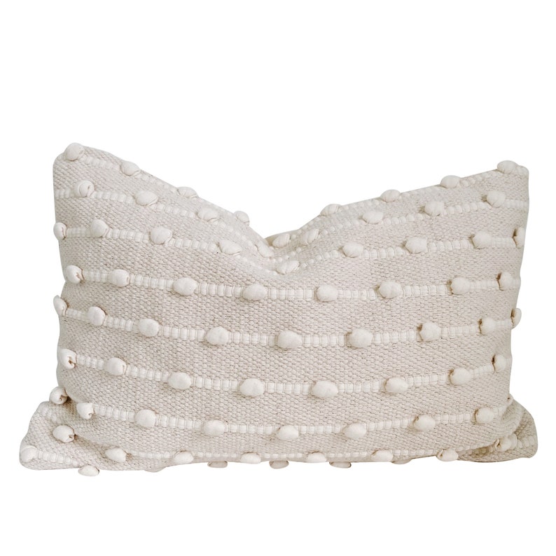 May include: A cream-coloured throw pillow with a textured woven design featuring small white pom-poms.