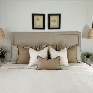 May include: A beige linen bed with a light brown headboard and a set of beige and brown throw pillows. Two framed botanical prints of palm trees hang above the headboard.