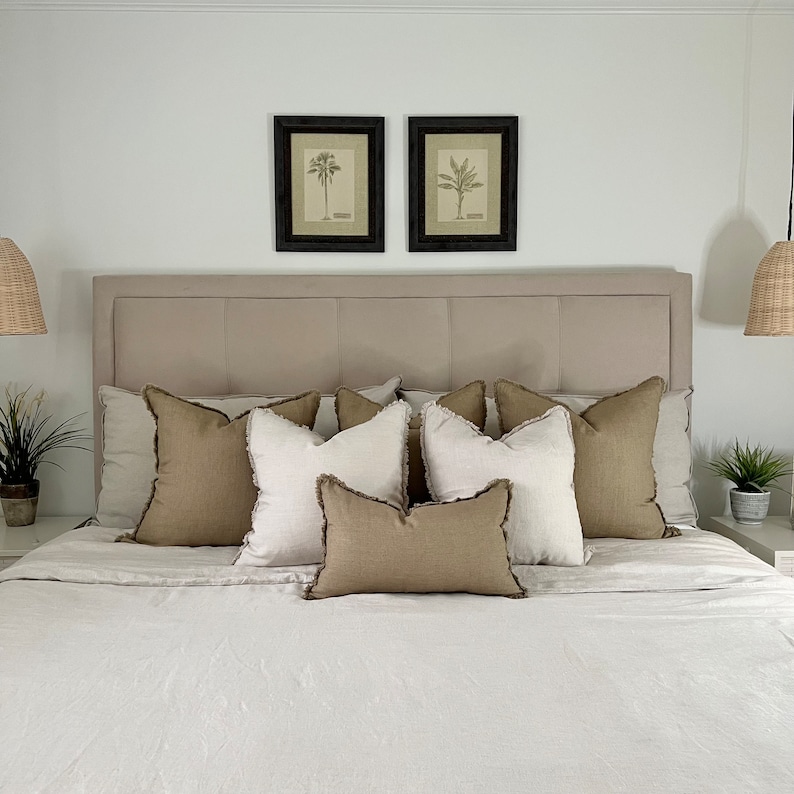 May include: A beige linen bed with a light brown headboard and a set of beige and brown throw pillows. Two framed botanical prints of palm trees hang above the headboard.